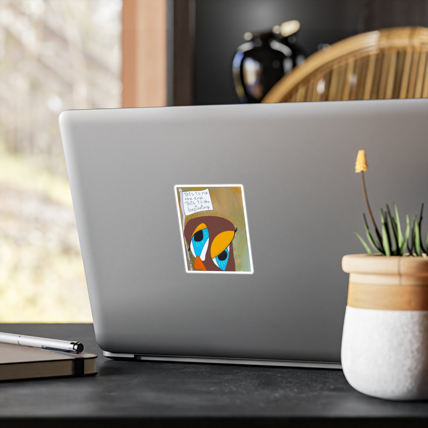 A silver laptop on a desk is decorated with the Printify "Vinyl Decal - Sobriety Gifts, This is Not The End," featuring colorful abstract art and handwritten text. Nearby are a notebook, pen, plant, and household items.