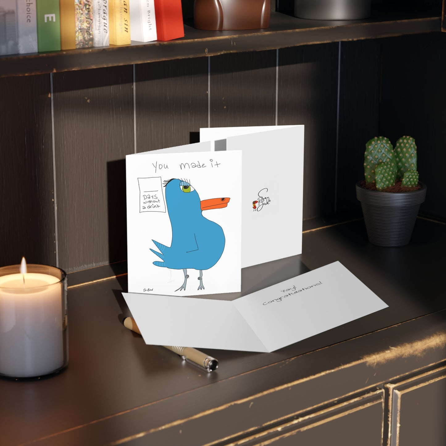 Printify’s Greeting Cards Recovery Celebration sits on a dark wooden shelf with a lit candle, pen, and two cacti. The main card shows a cartoon blue bird and “You made it”—an ideal milestone celebration card.