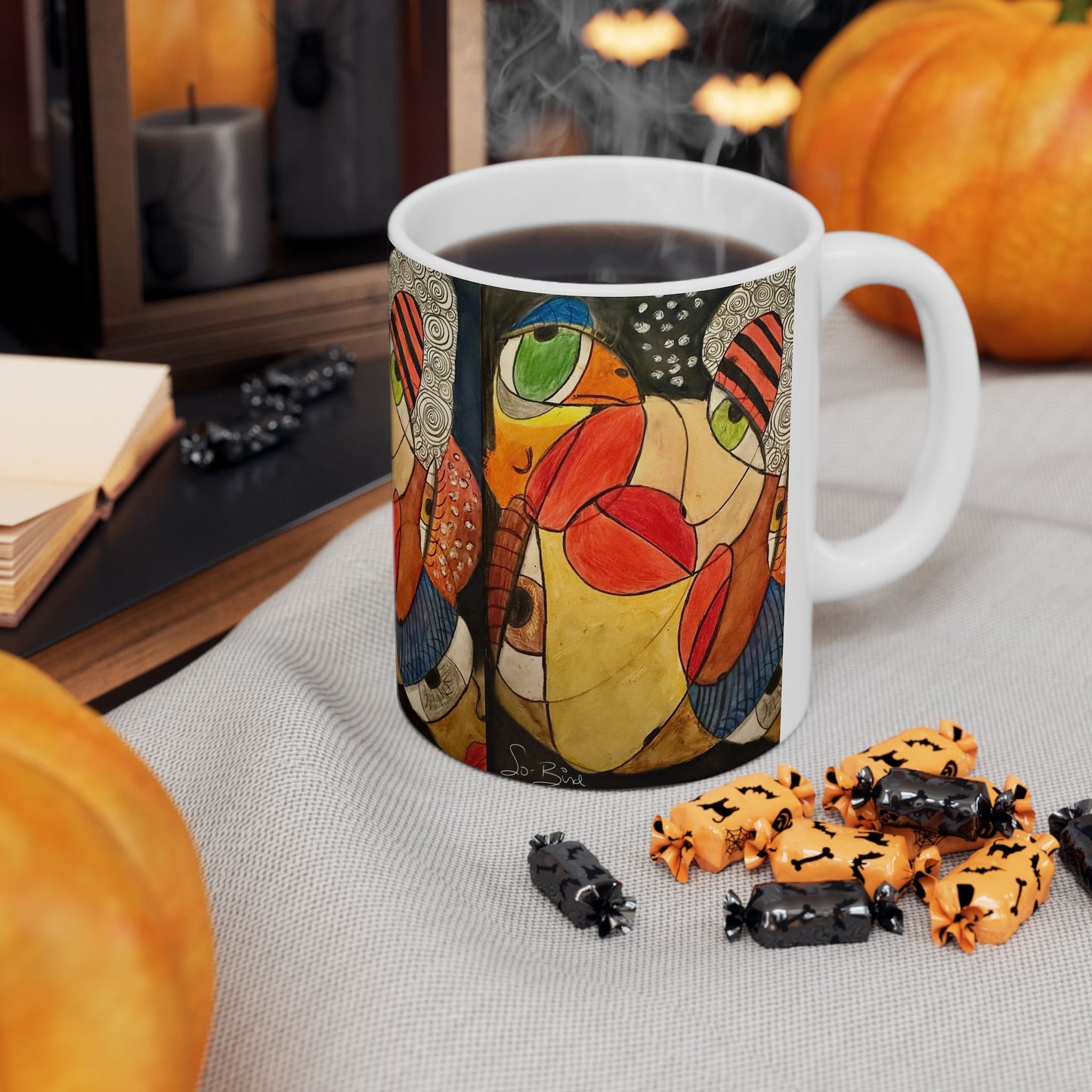 The Printify Ceramic Mug – It's a WE thing Recovery and Sober Gift – features a colorful abstract face and sits among Halloween candies, pumpkins, and a book. Steam rises from the mug, creating a cozy autumn vibe for those in recovery.