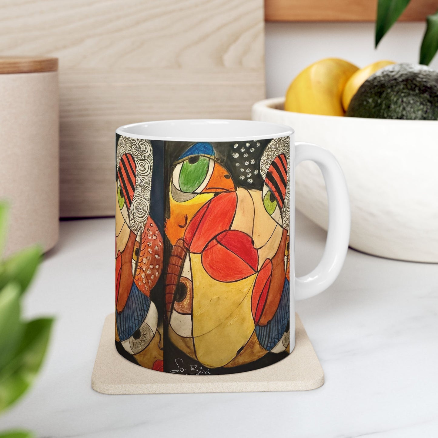 The Printify Ceramic Mug - It's a WE thing Recovery and Sober Gift, with its bold, colorful abstract design, sits on a kitchen counter—ideal for your morning coffee. In the background, a white bowl of fruit and potted plants add charm.