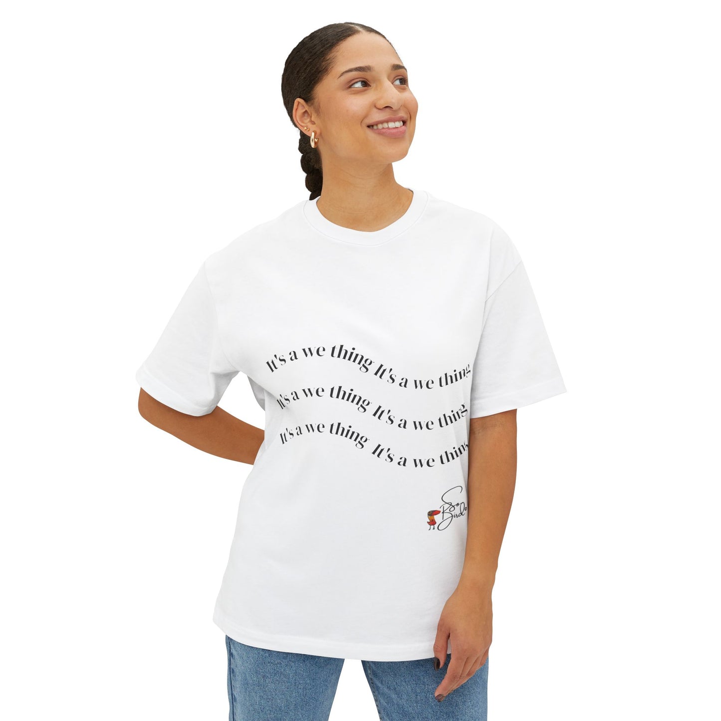 A person with tied-back hair smiles and looks to the side, wearing the Printify Recovery Support Unisex Tee featuring repeated “It’s a we thing” text and a small red and black graphic, paired with blue jeans for comfort.