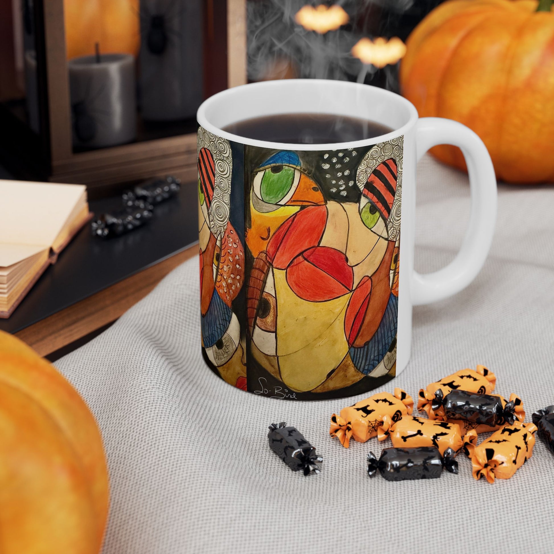 The Printify Ceramic Mug – It's a WE thing Recovery and Sober Gift – features a colorful abstract face and sits among Halloween candies, pumpkins, and a book. Steam rises from the mug, creating a cozy autumn vibe for those in recovery.