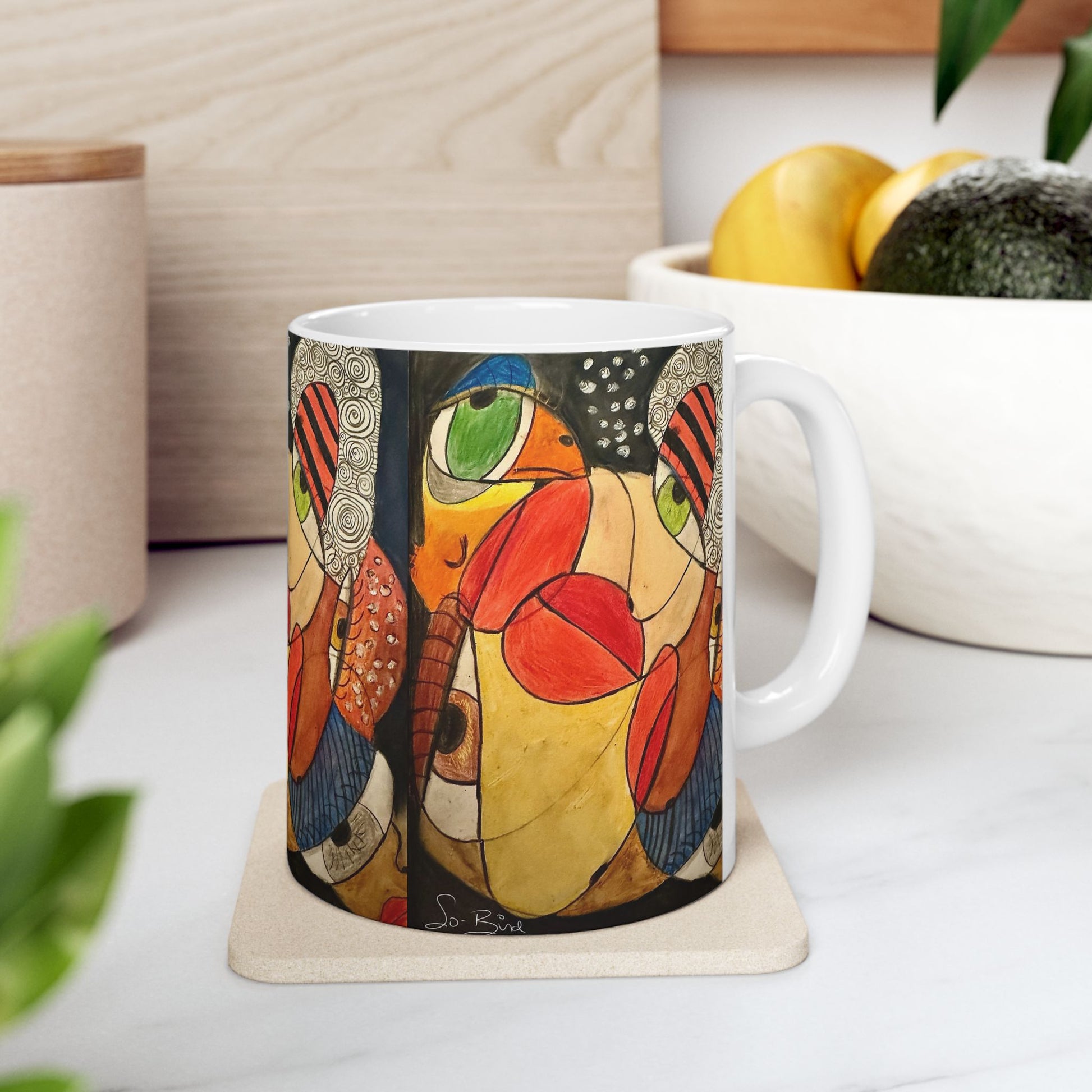 The Printify Ceramic Mug - It's a WE thing Recovery and Sober Gift, with its bold, colorful abstract design, sits on a kitchen counter—ideal for your morning coffee. In the background, a white bowl of fruit and potted plants add charm.